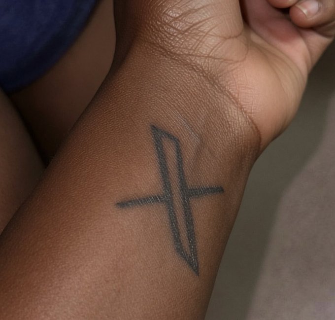 A close-up of a dark-skinned wrist displaying a black ink tattoo shaped like a stylized number '4' or lightning bolt symbol.