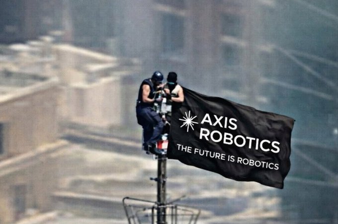 Two people sit on top of a structure holding a large black flag that reads 'AXIS ROBOTICS - THE FUTURE IS ROBOTICS' against a backdrop of a smoke-filled city skyline.