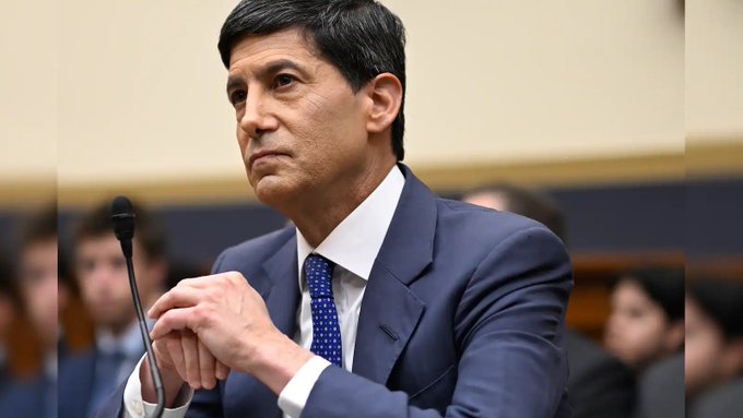 A man in a navy suit and blue patterned tie sits at a microphone in what appears to be a congressional or committee hearing, with blurred figures visible in the background.