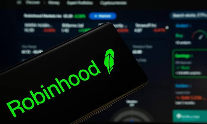 A smartphone displaying the green Robinhood logo and app icon in front of a blurred computer screen showing trading data with various stock or crypto metrics.