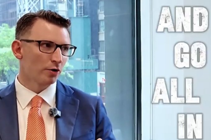 A bespectacled man in a blue suit and orange tie speaks during an interview, with the bold text "AND GO ALL IN" displayed beside him, visually reinforcing his declaration that he is fully committed to dedicating his life to Bitcoin.