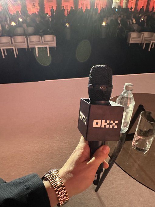 The image features a person holding a microphone with an OKX logo, possibly at a social gathering or event. The background shows several chairs arranged around tables, suggesting that this could be a conference or meeting setting. A water bottle can also be seen on one of the tables. The overall atmosphere appears to be relaxed and informal, as if the person is giving a speech or announcement during the event.
