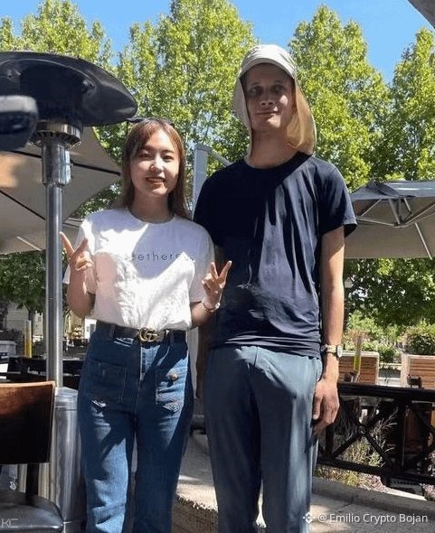 Two people pose outdoors at a cafe with patio heaters - an Asian woman in a white t-shirt and jeans making peace signs next to a tall blonde man in a navy shirt and bucket hat, with watermark '@_Emilio Crypto Bojan'.