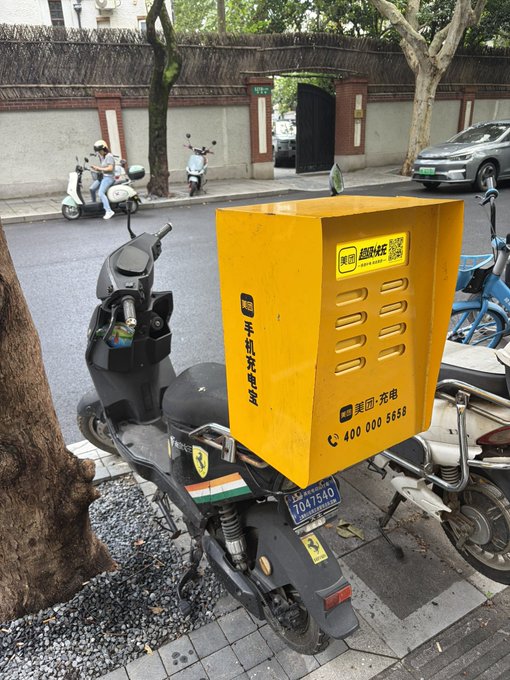 An electric scooter parked on a city street with a large yellow Meituan (美团) delivery box attached to the back, featuring the text '手机充电宝' and a customer service number.