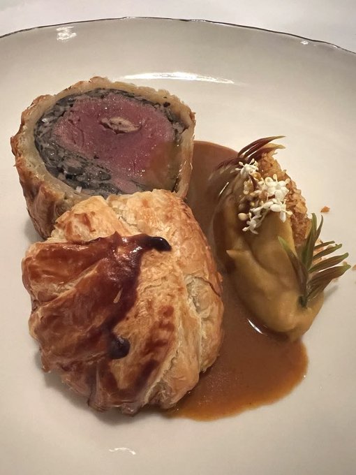 A fine-dining plated dish featuring a sliced Wellington-style beef tenderloin wrapped in pastry with a duxelles mushroom layer, accompanied by a golden pastry parcel and a garnish of white flowers and rosemary, served with a rich brown sauce.