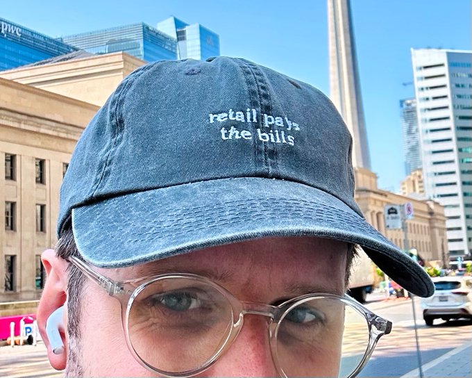 A close-up selfie of a man wearing clear-framed glasses and a denim baseball cap embroidered with 'retail pays the bills', photographed in an urban setting.