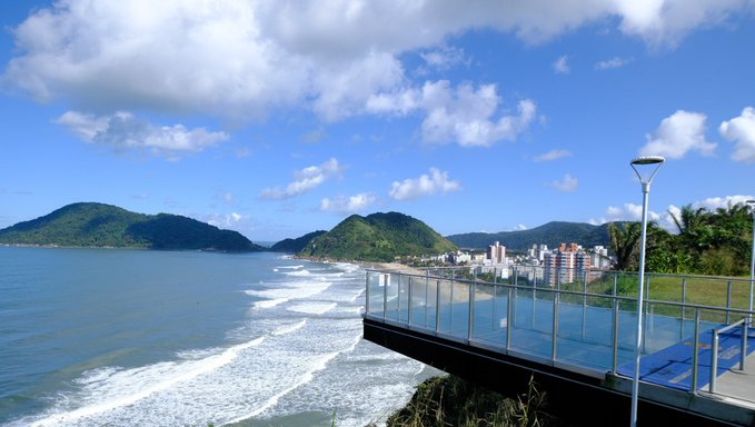 A coastal landscape featuring a glass-railed observation deck overlooking a blue ocean with waves crashing on the beach, with a city nestled at the base of green mountains under a partly cloudy sky.