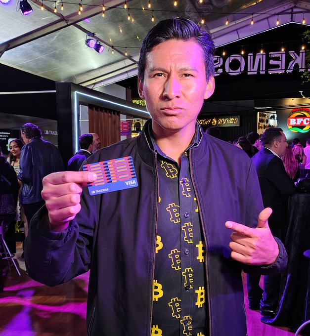 A man in a dark jacket and Bitcoin-patterned shirt holds up a VISA debit card at what appears to be a crypto event, with a partially visible 'KENIORS' sign and BFC logo in the background.