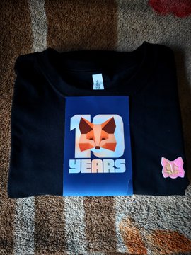 A folded black t-shirt is displayed on a striped carpet, featuring a blue graphic with a fox face and the text "10 YEARS," accompanied by a small pink butterfly sticker. The image showcases the MetaMask 10 Year Anniversary t-shirt swag mentioned in the post, celebrating the ambassador's recognition from the MetaMask ecosystem.