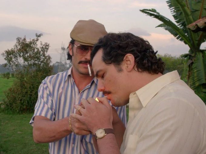 Two men stand outdoors near palm trees and greenery; one wears a striped shirt, sunglasses, and a tan flat cap while smoking a cigarette, while the other in a white shirt lights a cigarette, displaying a wristwatch and gold rings.