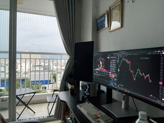 A home office setup featuring two monitors—one displaying trading charts and the other showing YouTube—with a keyboard, PC tower, and a balcony view of a city.