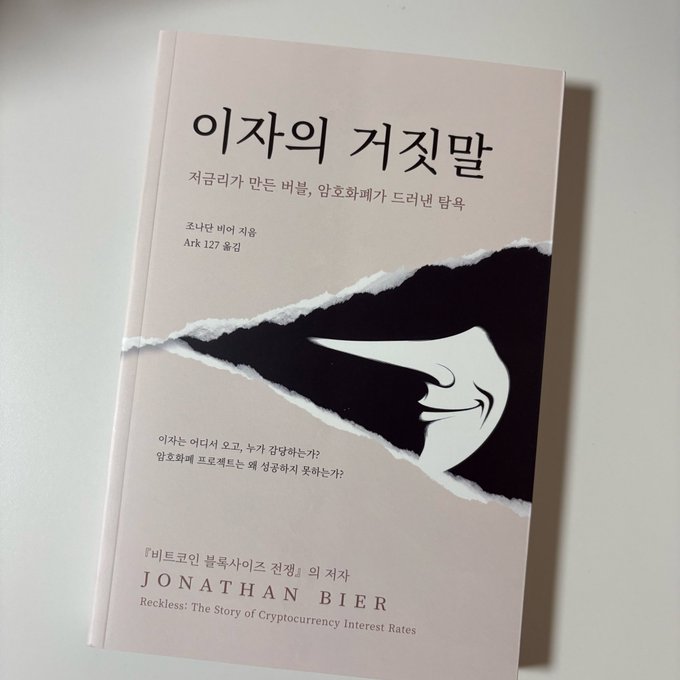 A book titled '이자의 거짓말' (The Lie of Interest) by Jonathan Bier, with English subtitle 'Reckless: The Story of Cryptocurrency Interest Rates' and translator Ark, featuring a torn paper design with a pointing finger.
