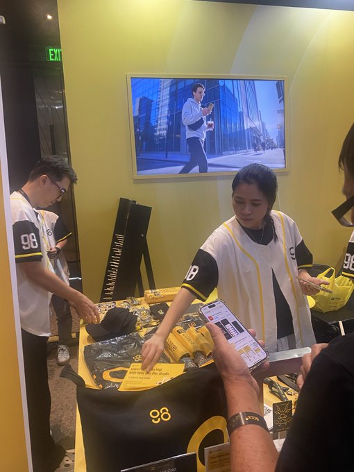 An indoor promotional event or merchandise booth where staff wearing '98' jerseys are handing out merchandise, including black and yellow hats and t-shirts.