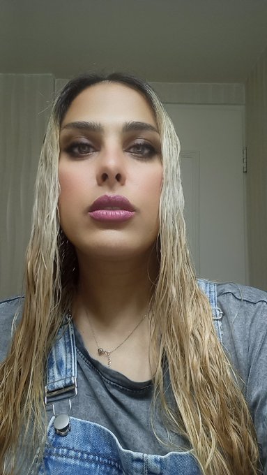 A close-up selfie of a person with long blonde hair, wearing a denim jacket over a gray top and a delicate necklace.