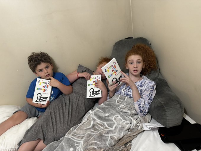 Three children lie together on a bed with gray bedding, each holding a book titled 'things not enough' featuring a swimmer illustration on the cover.