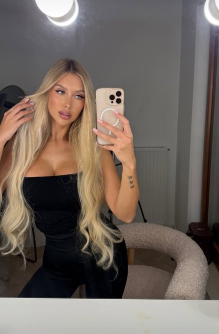 A blonde woman with long wavy hair takes a mirror selfie in a black strapless top, holding a phone with a pop socket.
