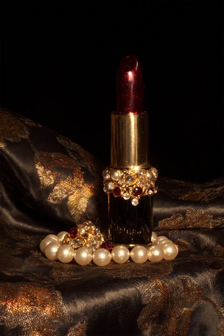 An elegant photograph of a red lipstick tube adorned with jewels, resting on golden pearl necklaces against dark satin fabric with gold patterns.
