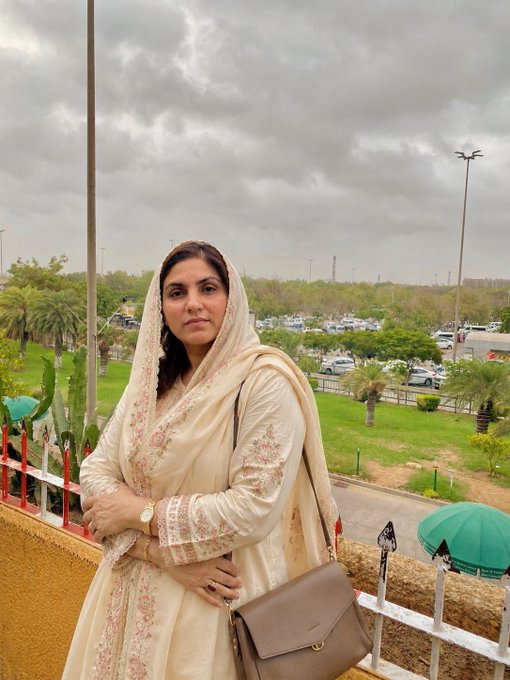 A woman in a cream-colored traditional dress with a matching embroidered dupatta stands on a balcony, holding a brown handbag, with a parking lot and cloudy sky visible behind her.