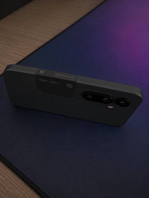 The image features a black smartphone lying face down on a blue surface, such as a mouse pad or desk. The phone appears to be turned off with its screen facing upwards. In the background, there are two texts written in white that read "my wife just saw me carrying an iPhone and a @solanamobile she thinks I have a second life". This post seems to be about someone's experience of using their smartphone while being watched by another person or a character from the @solana game. The focus on this particular image suggests that it might be related to the gaming community, with the presence of the @solana game in the background.