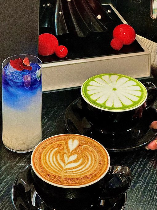 A photograph of two elaborately prepared coffees on a dark table—one latte with a heart/leaf design and a green matcha with a flower design—alongside a striking blue and white layered drink with rose petals.
