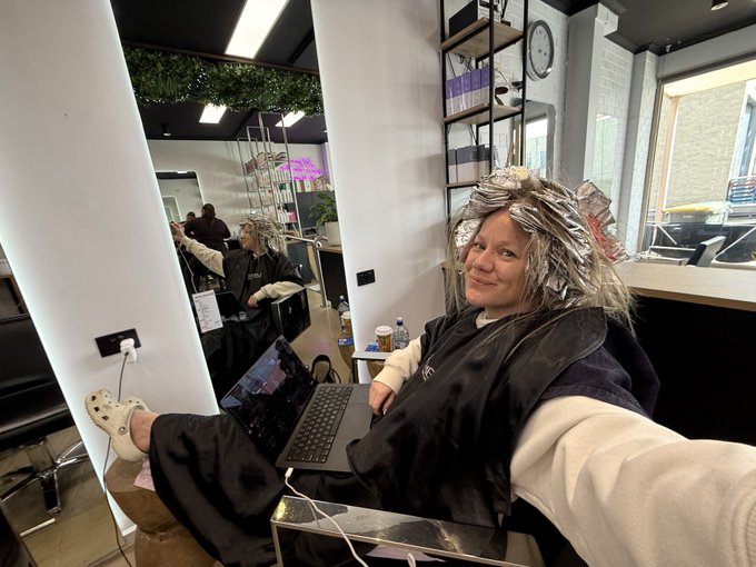 A selfie of a woman at a hair salon with foils in her hair, wearing a black cape, smiling while holding up her feet in Crocs on a counter next to an open laptop.