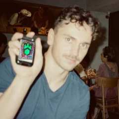 A man with curly hair smiles while holding up a small handheld gaming device displaying a green pixelated game on its screen, appearing to promote or relate to the "$25 SOL GIVEAWAY" mentioned in the accompanying post. The casual indoor setting and playful pose suggest he's engaging with the crypto giveaway promotion.