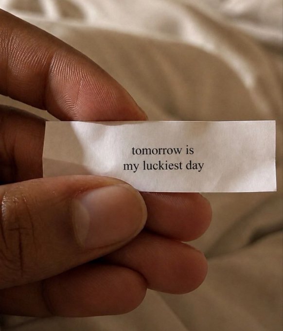 A hand holds up a fortune cookie slip reading "tomorrow is my luckiest day," a personal affirmation being "claimed" as a hopeful declaration of good fortune. The intimate, warm setting reinforces the message of embracing optimism and manifesting luck in the day ahead.