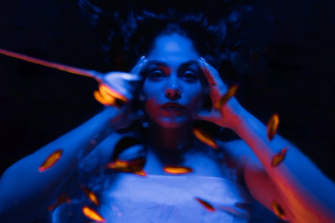 A woman with glowing blue skin and red lipstick holds her hands to her temples, surrounded by floating orange flame-like shapes against a dark background.