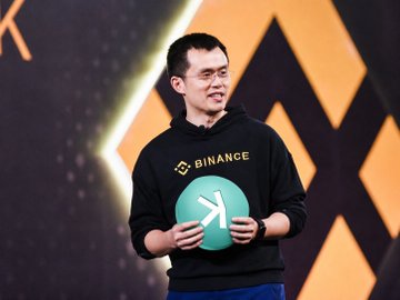 A man wearing a black Binance hoodie stands on stage holding a teal balloon with a white logo, appearing to speak at a Binance event with a geometric background.