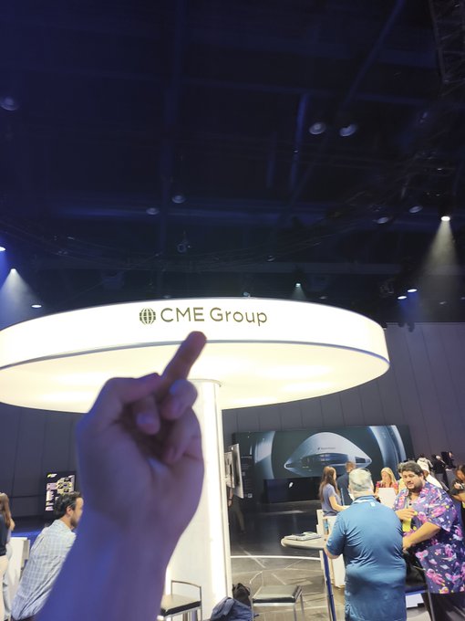 A photo taken at an event showing a hand giving a middle finger gesture toward the CME Group booth, with attendees visible in the background.