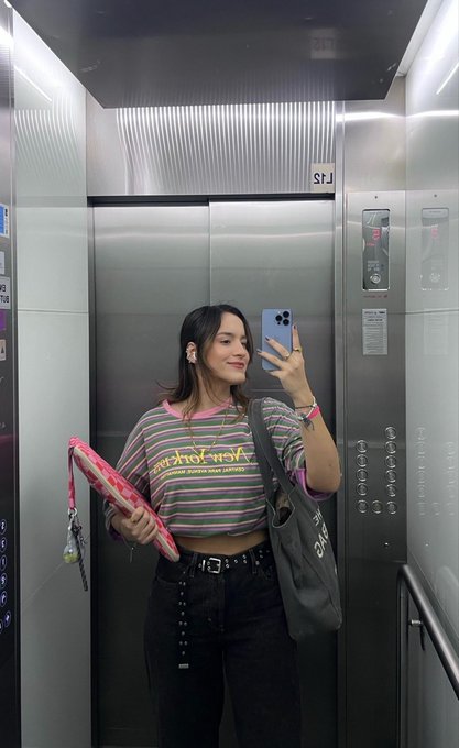 A young woman takes a mirror selfie in an elevator wearing a striped 'world news' shirt, holding a pink patterned bag and a gray tote bag with earbuds in.