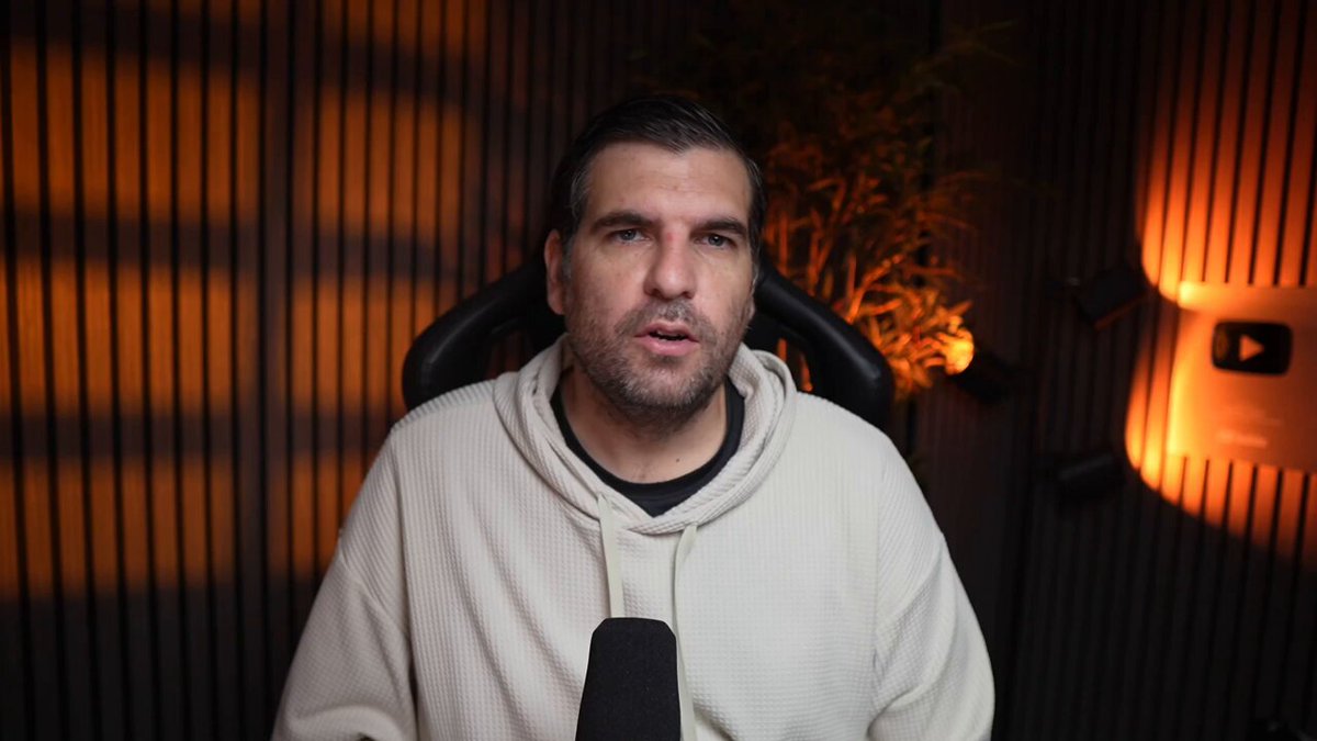 In this image, there's an adult male wearing a white hoodie who appears to be a game host or interviewer with a microphone in front of him. He seems to be engaged in a conversation, which is possibly related to gaming or another topic. The background features a combination of orange and black colors, creating a striking visual effect. The text above the image reads: "Hedera just built a $200B corporate trap Billions of dollars remain trapped inside legacy corporate loyalty systems, creating a major financial headache and unredeemed liabilities on corporate balance sheets". This message highlights an important issue in the world of gaming or entertainment where companies may exploit their users' personal information for financial gains.