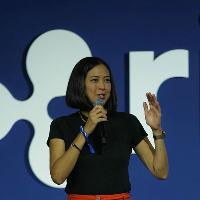 A woman with short dark hair holds a microphone and gestures while speaking, standing in front of a blue backdrop with white letters.