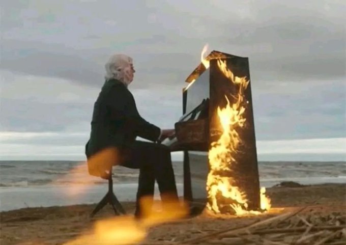 An older man with white hair plays an upright piano that is engulfed in flames on a sandy beach with the ocean in the background.