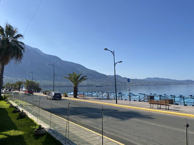 The image shows a street with palm trees lining both sides, leading towards a body of water that has mountains in the background. The road appears empty at the moment, but there are several cars parked along its length. A bench can be seen on one side of the street, and a person is standing near it. In addition to these elements, there's also a post with text that reads "Gm $Dog from Kalamata We are going to the moon very soon". This message seems to be related to an event or promotion happening in the area.
