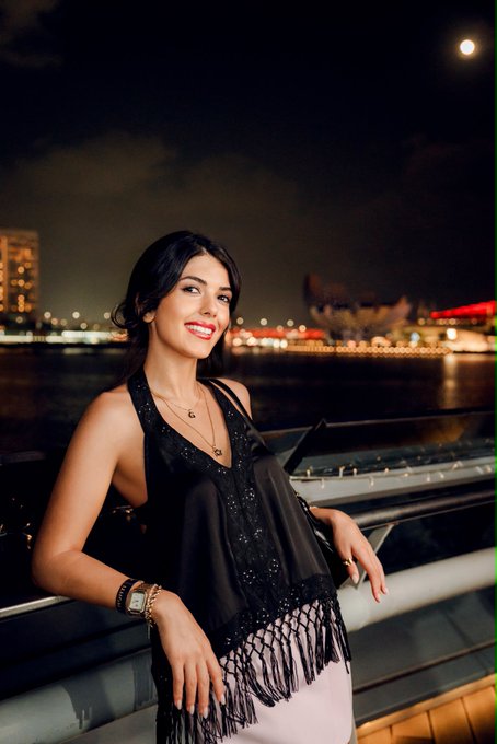 A portrait of a smiling woman with dark hair wearing a black fringed top, posed at night with an illuminated city skyline across the water and a full moon visible in the sky.