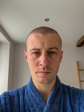A selfie of a bald man with a serious expression, wrapped in a blue towel or robe, photographed indoors.