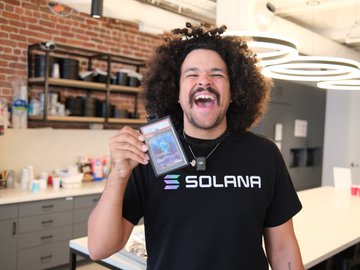 A man with curly hair laughing joyfully while holding up a card, wearing a black t-shirt with 'SOLANA' printed on it.
