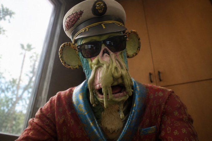 A person in heavy green monster makeup with glasses and round earrings wears a captain's hat and a patterned robe, sitting near a window.