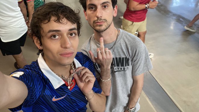 A selfie shows two young men, one in a blue Nike polo with a heart necklace, the other in a gray 'HAWK' t-shirt making a middle finger gesture, standing in what appears to be a crowded public space.