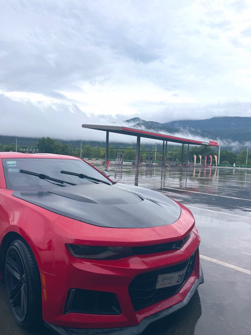 A red Chevrolet Camaro ZL1 sports car with a black hood parked at a gas station on a rainy day, with mist-covered mountains in the background.