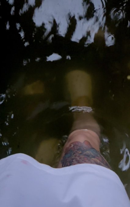 A first-person view looking down at a tattooed leg and foot submerged in murky water, with a beaded anklet visible around the ankle.