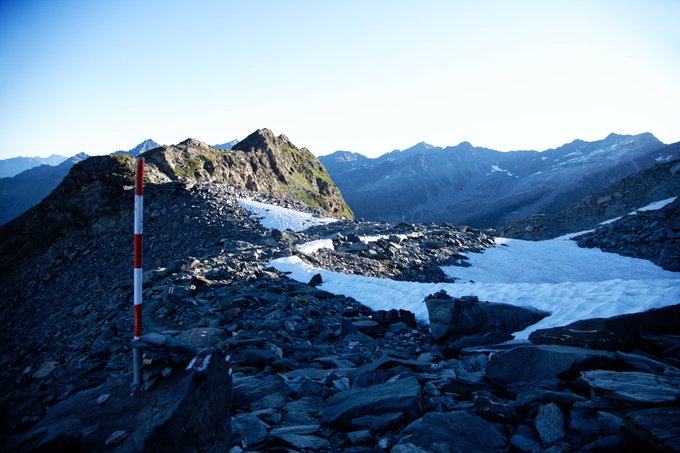 A scenic mountain landscape with rocky terrain, patches of snow, and a red-and-white striped marker pole under a clear blue sky.
