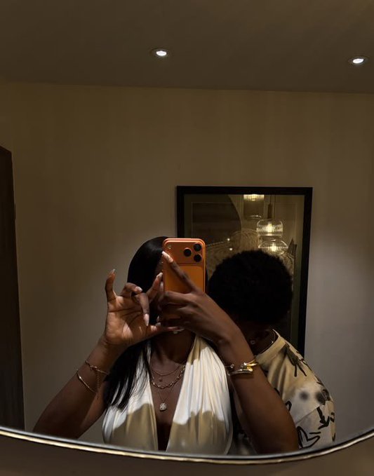 The image shows a couple taking a selfie together, with one person holding up their phone to take the picture while the other person looks at the reflection in the mirror. The background features a white wall and a mirror that reflects the room's interior lights. There is also an orange object on the floor near the mirror.