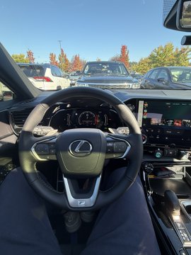 Interior view from the driver's seat of a Lexus vehicle, showing the steering wheel with the Lexus logo, digital dashboard, and a parking lot visible through the windshield.