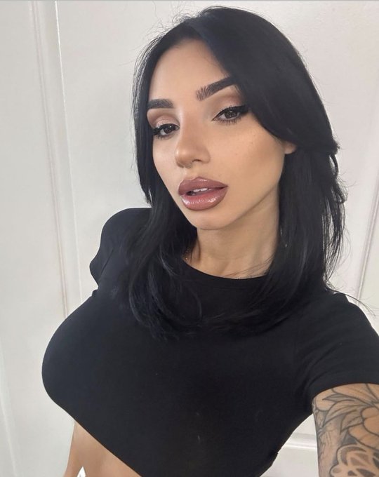 A selfie of a woman with long black hair wearing a black t-shirt, featuring prominent makeup and a tattoo on her arm.