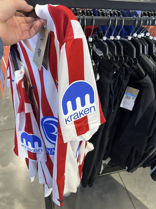 A hand holding up a red and white striped soccer jersey featuring the "Kraken" cryptocurrency logo on the sleeve, displayed on a rack alongside other merchandise.