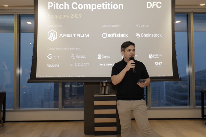 A man in a black shirt and khaki pants speaks into a microphone while standing in front of a presentation screen titled "Pitch Competition Singapore 2026," featuring sponsor logos including Arbitrum, softstack, and Chainstack.