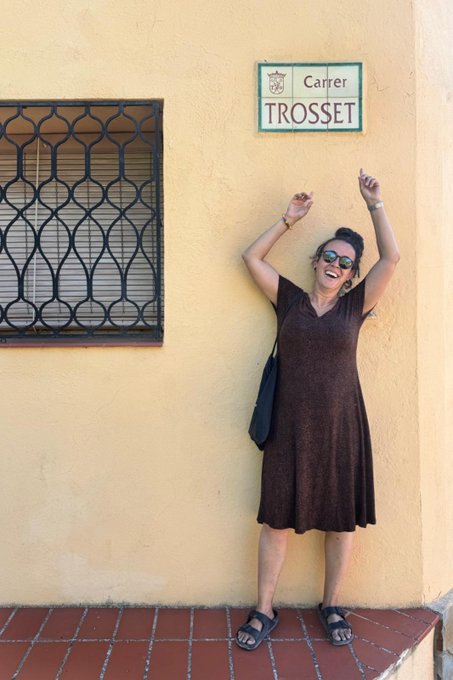 A woman in a brown dress poses joyfully with arms raised in front of a yellow wall beneath a street sign reading "Carrer Trosset," celebrating her discovery of a piece from her AB500 art collection in the small village of Tivissa near Tarragona, Spain.
