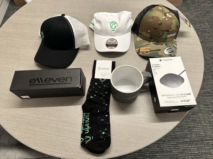 The image shows an array of Uphold-branded merchandise and tech items arranged on a table, including three caps (black trucker, white, and camo), a black "el leven" box, Uphold constellation-patterned socks, a ceramic mug, and a Mophie 15W wireless charging pad, all in their original packaging. These items appear to be the giveaway prizes for the @Upholdinc 40K subscriber celebration mentioned in the post, likely featuring products that the YouTuber has reviewed or uses.